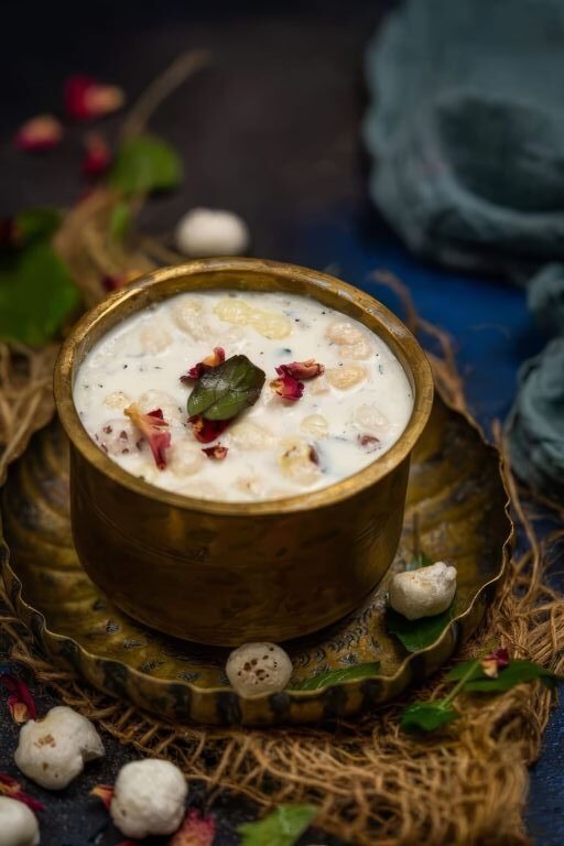 A brass bowl filled with Panchamrut, a traditional Indian mixture, is garnished with rose petals and a fresh green leaf. The bowl is placed on an ornate brass plate, with some puffed lotus seeds scattered around. The background features rustic and natural elements, including dried twigs and leaves, creating a traditional and earthy ambiance.
