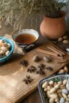 A rustic wooden table is adorned with various spices and nuts. The center features a wooden cutting board with star anise, nutmeg, and cinnamon sticks arranged on it. Surrounding the board are ceramic bowls containing almonds and pistachios. A cup of tea in a decorative bowl sits nearby. In the background, a terracotta pot holds dried herbs. The scene is warmly lit, creating a cozy and inviting atmosphere.